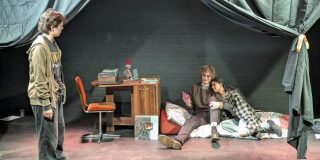 A photo of three cast members on set. Two are lying down whilst one is standing and speaking to them.