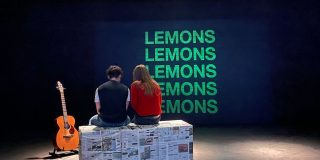 Two actors sit on a bench, next to a guitar.