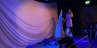 Four actors gesticulate to each other in front of a large white sheet. The photo is taken at a side angle.