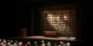 Image of Oxford Playhouse's 'The Gang of Three' set, consisting of a large bookshelf, sofa, and the audience below the stage.
